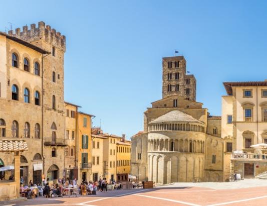 Piazza medievale soleggiata con chiesa romanica e caffè all'aperto, atmosfera storica e rilassata.