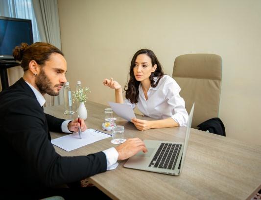 Due persone discutono in ufficio con un laptop e documenti su un tavolo.