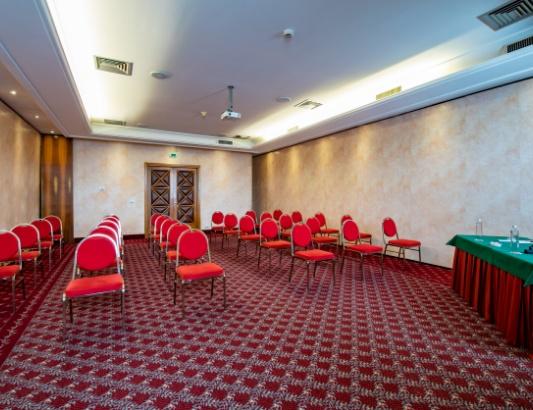 Sala conferenze con sedie rosse e tavolo verde su tappeto decorato.