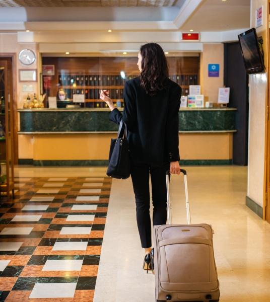 Woman with suitcase enters hotel, reception in the background.