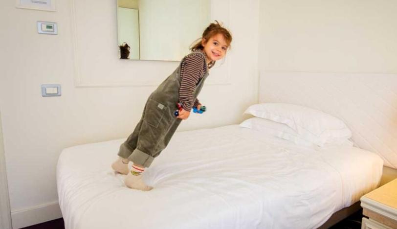 Bambina sorridente gioca saltando su un letto in una stanza luminosa.