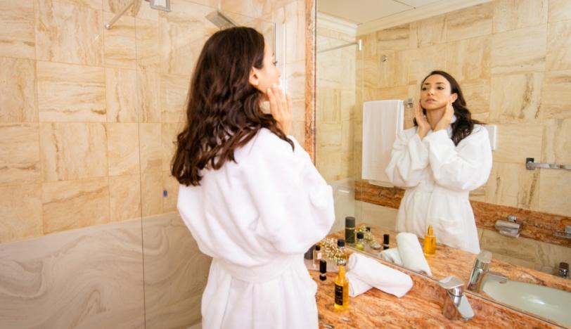 Donna in accappatoio applica crema viso davanti allo specchio del bagno elegante.